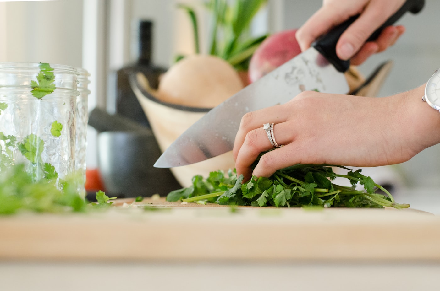 Hands chopping a bundle of cilantro on a wooden board, a chef’s knife mid-cut.