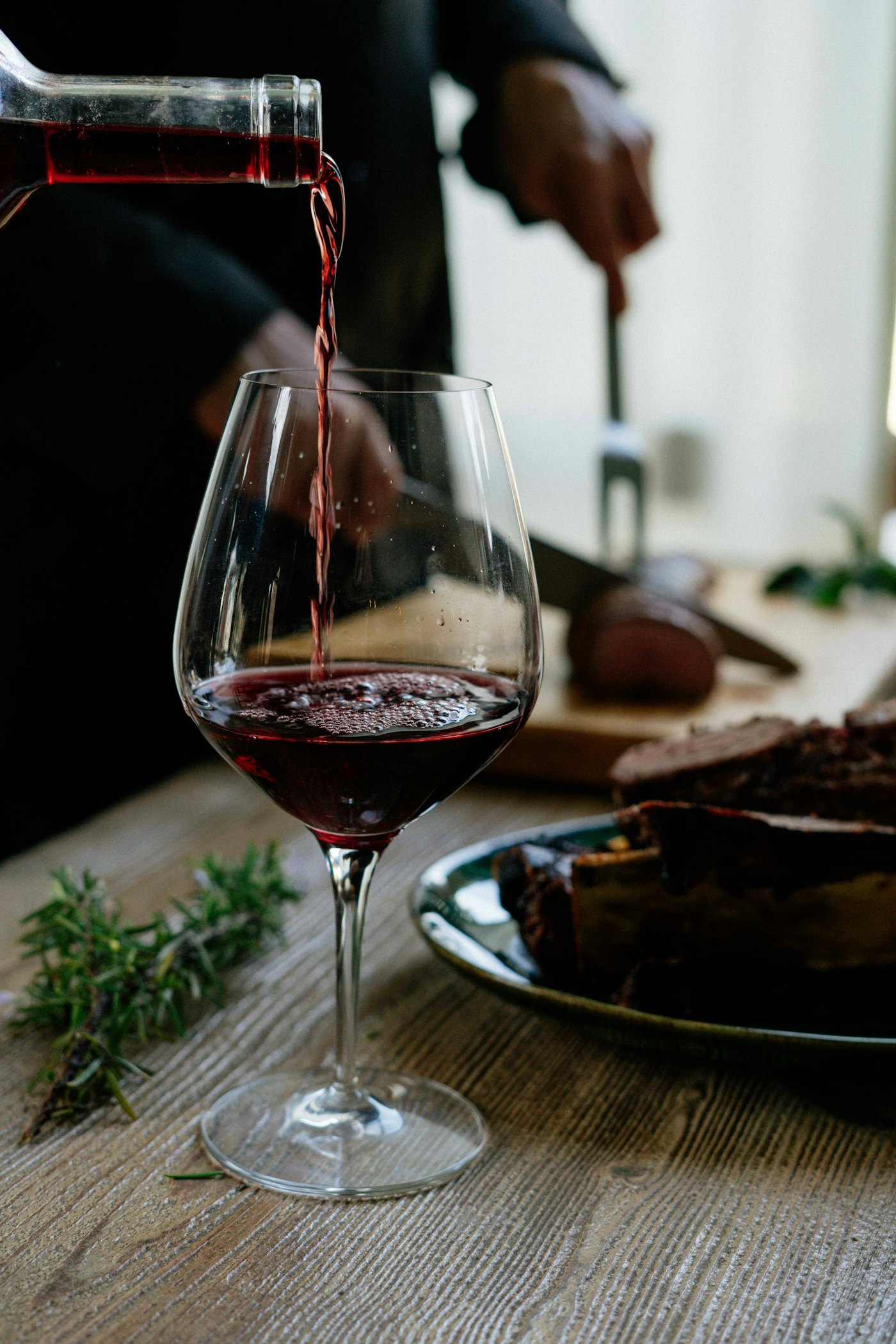 Red wine pouring from a bottle into a glass on a wooden table, with a plate and carving in the background.