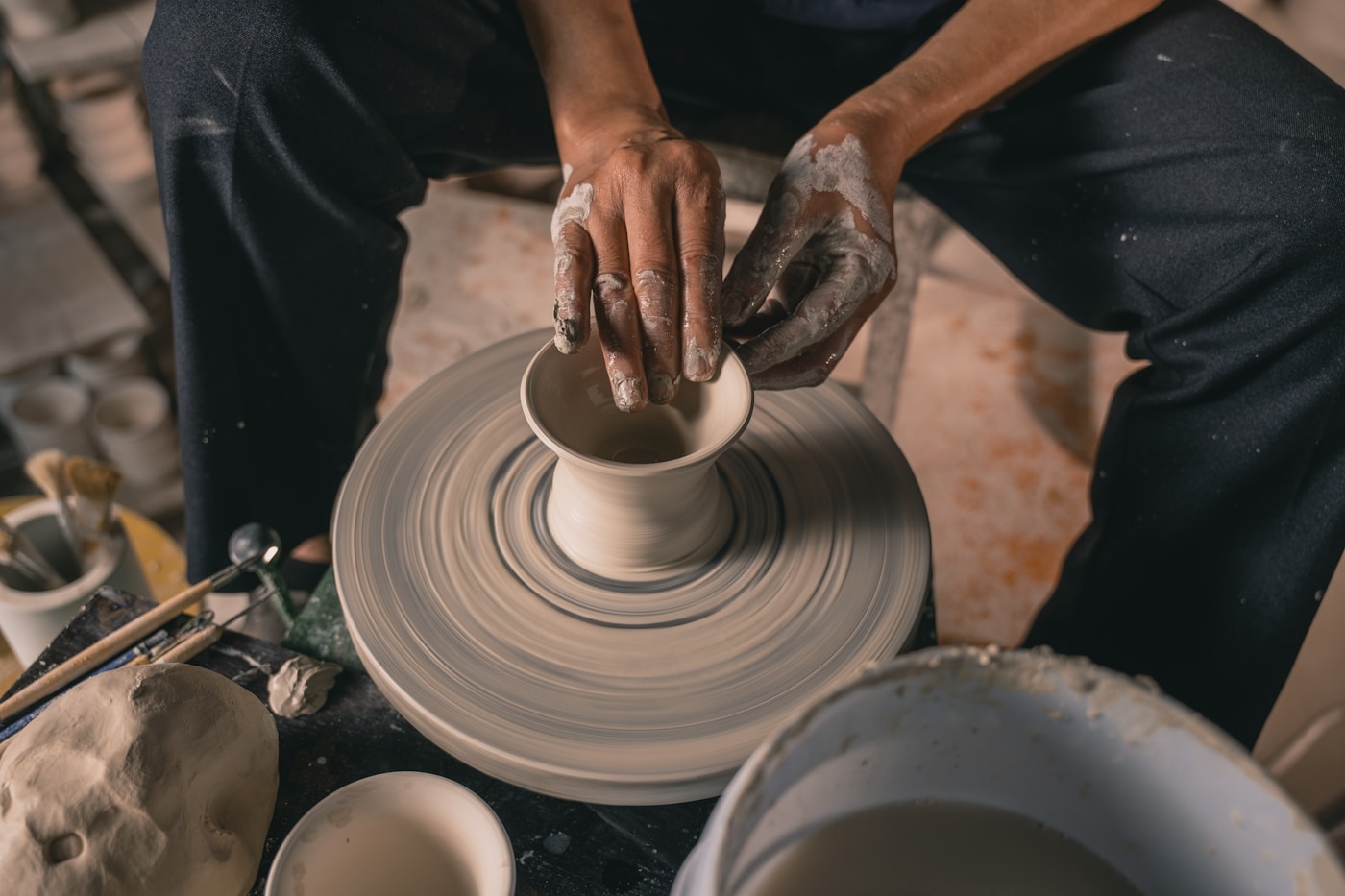 Clay-covered hands shaping a small vessel on a pottery wheel.