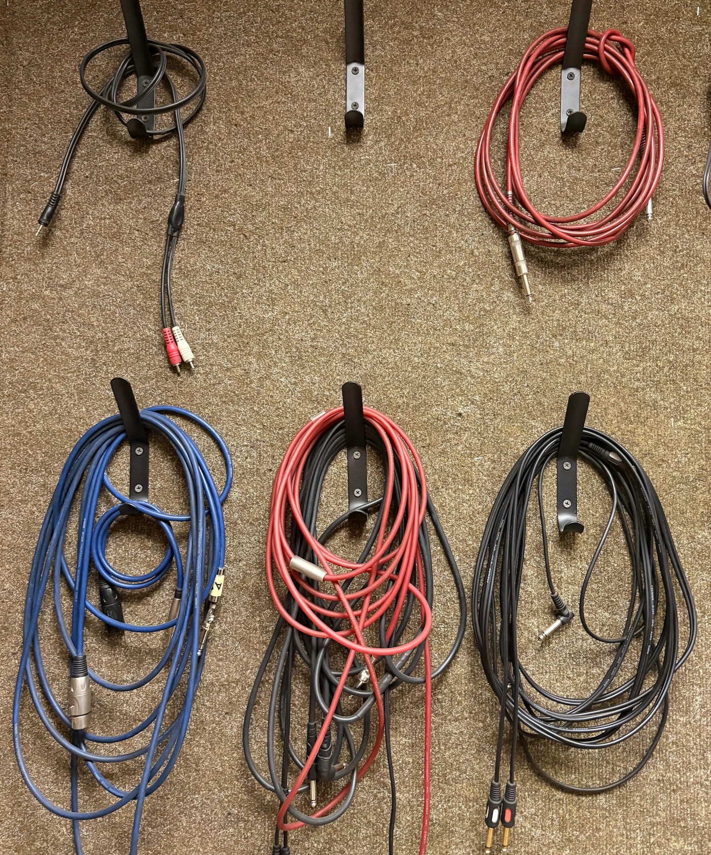 Coiled red, blue, and black audio cables hanging from metal hooks on a beige carpeted wall.