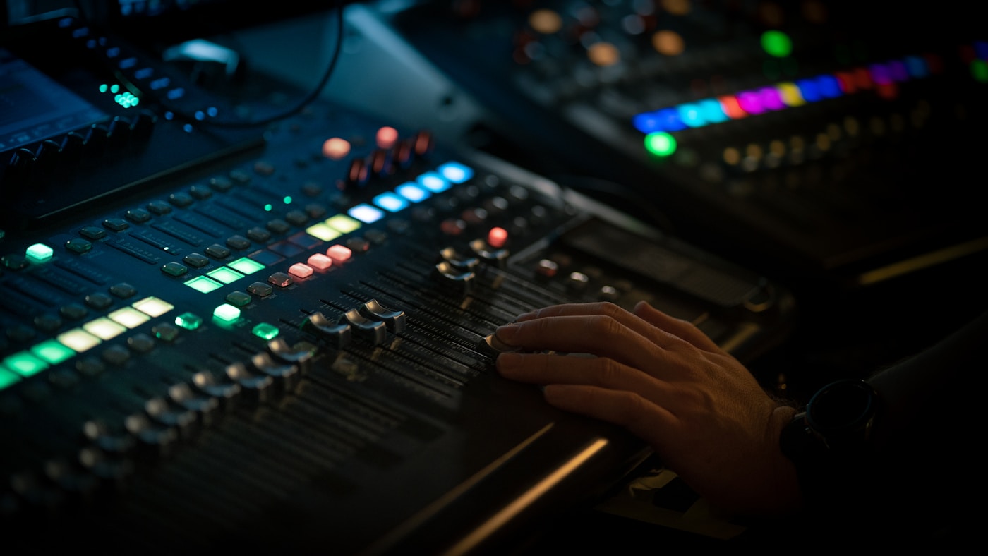 A hand resting on the faders of a mixing console, with coral, green, and blue button lights nearby.