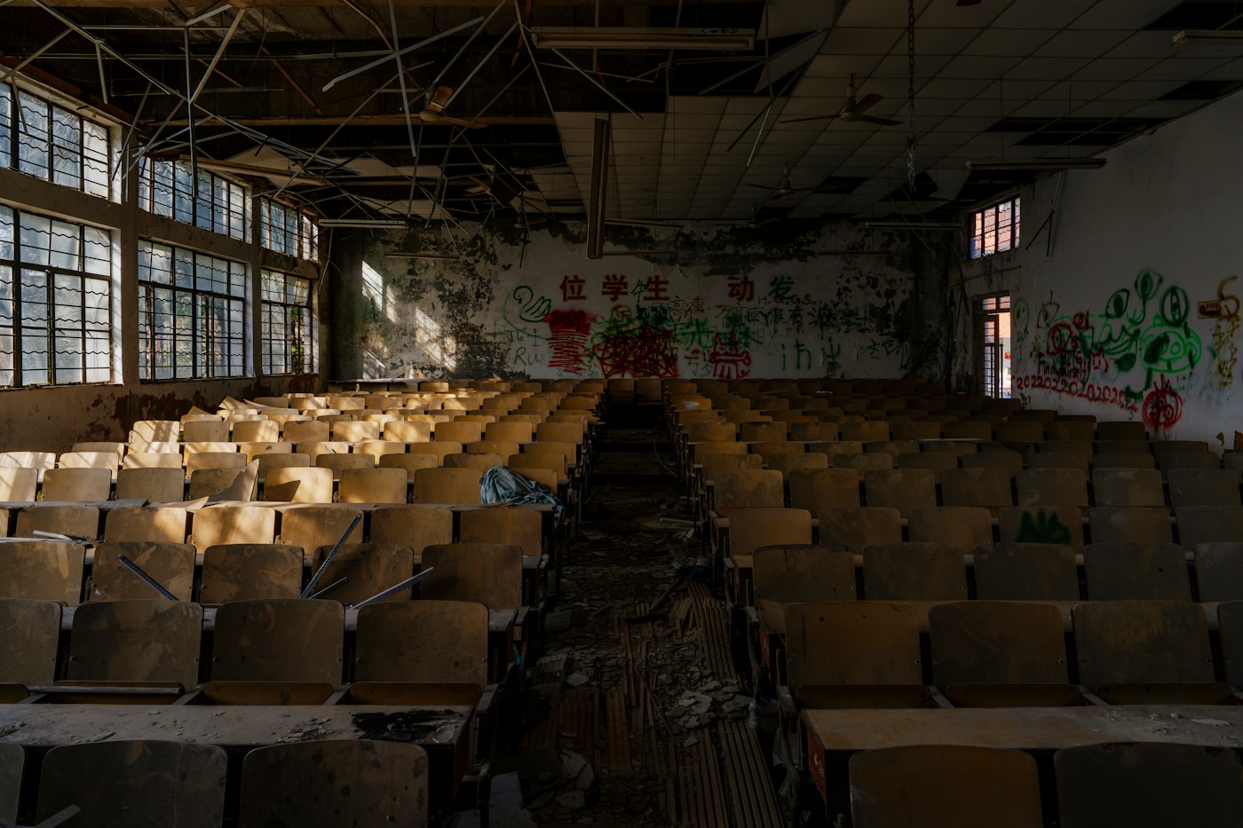 Rows of empty wooden desks in a unused, sunlit hall with damaged ceiling and graffiti on the far wall.