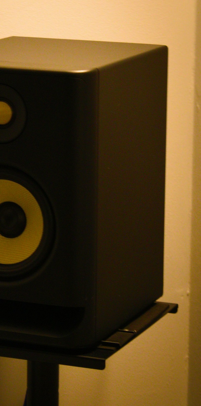 Close view of a black studio monitor on a stand, showing a yellow woofer cone and a plain wall behind it.